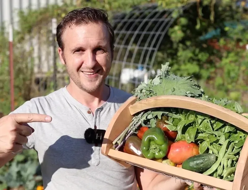 Paul Dysinger holding a wooden basket filled with freshly harvested peppers, kale, basil, cucumbers, and green beans